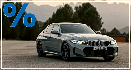 Fahrzeug: Eine BMW Limousine (vermutlich ein 3er Modell) in einer grauen oder hellblauen Lackierung.  Hintergrund: Das Auto steht auf einem leeren Park- oder Flugplatz, umgeben von Natur und Bergen im Hintergrund bei Dämmerung oder mildem Licht. Hohe Bäume sind ebenfalls sichtbar.  Aktionshinweis: Großes, blaues Prozentzeichen (%) in der oberen linken Ecke.  Kennzeichen: M SE 1466.