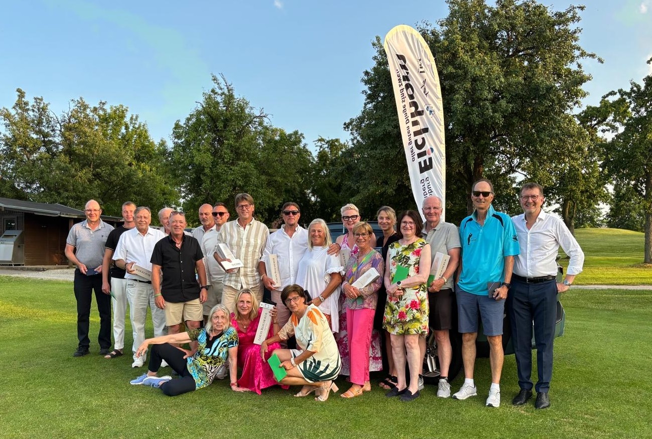 Eine große Gruppe von rund 20 Erwachsenen, Männer und Frauen, posiert am Abend auf einem Rasen, vermutlich nach einem Golf-Event. Einige Personen halten Trophäen oder Urkunden. Im Hintergrund steht eine weiße Werbefahne mit dem 'Eichhorn'-Logo. Die Stimmung ist locker und fröhlich, der Himmel ist in sanften Abendfarben gehalten