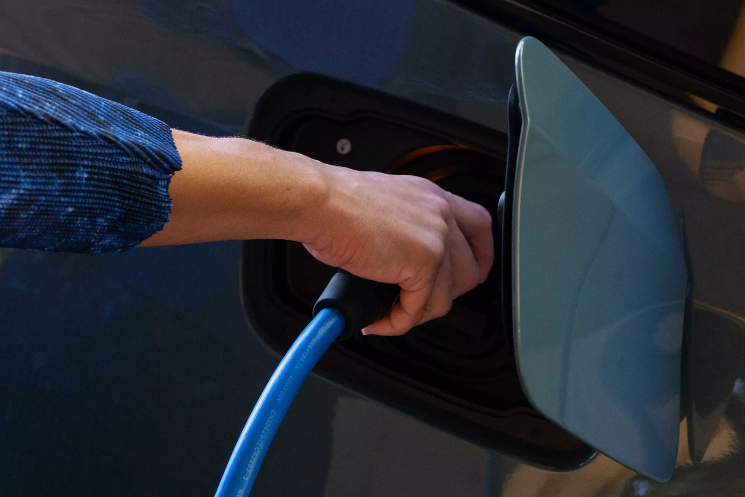 Eine Hand in einem blauen Ärmel hält ein blaues Ladekabel, das in den Ladeanschluss eines Autos gesteckt wird, der sich seitlich am Fahrzeug befindet. Der Hintergrund ist dunkel und unscharf.