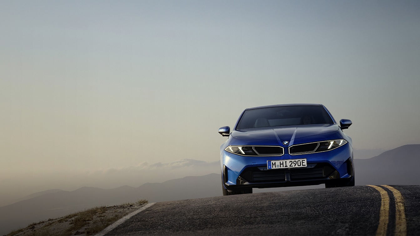 Neuer vollelektrischer BMW i3 in leuchtend blauer Lackierung aus der Frontperspektive auf einer Bergstraße. Die markante BMW Niere, die schmalen LED-Scheinwerfer und die sportlich gestaltete Frontpartie stehen im Mittelpunkt. Im Hintergrund erstreckt sich eine weitläufige Berglandschaft im sanften Licht der Abenddämmerung.