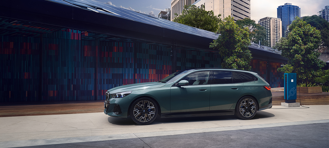 BMW i5 Touring in Seitenansicht an einer Ladestation vor moderner Stadtkulisse.