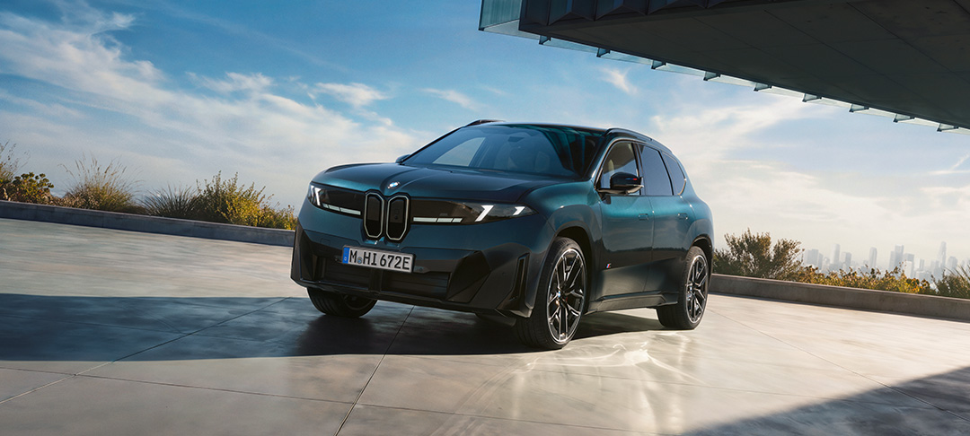 Neuer vollelektrischer BMW iX4 in dunkelgrüner Lackierung aus der vorderen Seitenperspektive. Das SUV-Coupé steht vor moderner Architektur und beeindruckt mit markanter BMW Niere, schmalen LED-Scheinwerfern und sportlichen Leichtmetallrädern.