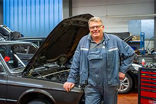 Ein lächelnder Mechaniker in blauer Arbeitskleidung steht in einer Werkstatt neben einem Auto mit geöffneter Motorhaube.