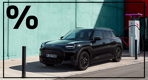 Ein schwarzer, kompakter Elektro-SUV, der vor einer helltürkisen Sichtbetonwand parkt. Das Fahrzeug wirkt robust und modern. Es ist gerade über ein schwarzes Kabel an eine weiße „Ionity“-Ladesäule angeschlossen. Ein großer Teil des Bildes liegt im harten Schatten, während das Heck des Wagens von der Sonne angestrahlt wird. Oben links prangt ein großes, fettes Prozentzeichen.