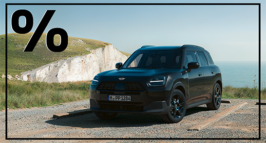 Das Bild zeigt einen schwarzen MINI Countryman C, der auf einem hellen Schotterparkplatz in einer weitläufigen Küstenlandschaft steht. Das Fahrzeug ist schräg von vorne aufgenommen, sodass die bullige Front und die linke Seite des SUV gut sichtbar sind. Im Hintergrund erhebt sich links eine markante, helle Steilküste mit grünen Grasflächen, während zur rechten Seite hin der Blick auf das ruhige, blaue Meer unter einem klaren, hellen Himmel freigegeben wird. Das Auto trägt das Kennzeichen „M PP 1284“ und zeichnet sich durch moderne LED-Scheinwerfer sowie dunkle Felgen aus. Markant ist zudem ein großes, fettes Prozentzeichen in der oberen linken Bildecke, und das gesamte Motiv wird von einem dünnen, schwarzen Rahmen umschlossen.