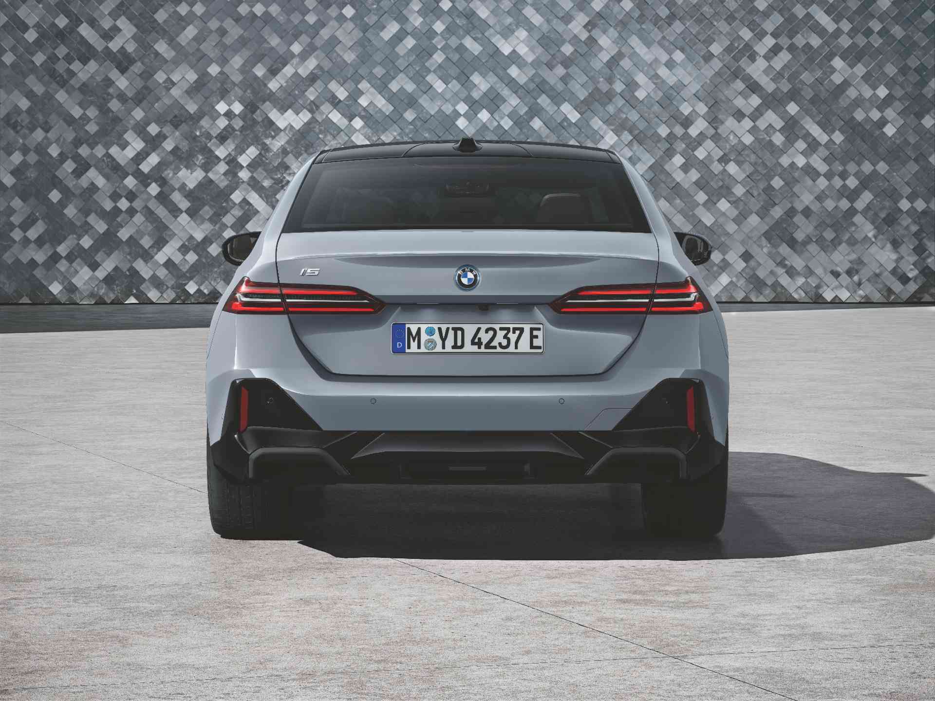 Ein silbergraues Auto steht auf einem hellgrauen gepflasterten Bereich vor einer Wand mit einem grauen, geometrischen Fliesenmuster. Das Auto hat schwarze Akzente und Felgen. Es ist leicht nach links gedreht, sodass die Vorderseite und die Fahrerseite zu sehen sind.