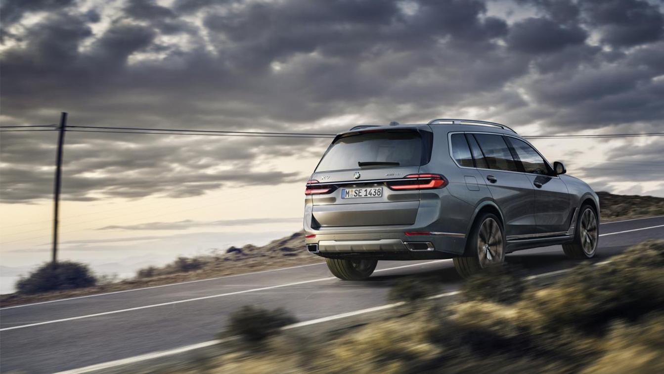 Ein seitlich-hinten aufgenommenes Bild eines grauen BMW X7, der auf einer leeren Straße bei Dämmerung oder Bewölkung fährt. Das Fahrzeug bewegt sich weg vom Betrachter, seine Rückleuchten und die Heckpartie sind gut sichtbar. Im Hintergrund ist eine weite, offene Landschaft mit einem bewölkten Himmel und einer Andeutung von Sonnenuntergang oder Aufgang zu sehen. Links im Bild steht ein Strommast oder eine Telefonleitung. Das Kennzeichen des Fahrzeugs lautet "M SE 1438".  Über dem Bild ist ein Textoverlay mit folgenden Informationen: WLTP Energieverbrauch kombiniert: 10,6 l/100 km (WLTP); CO2-Emissionen kombiniert: 240 g/km (WLTP); CO2-Klasse: G; Leistung: 280 kW (381 PS); Hubraum: 2.998 cm³; Kraftstoff: Benzin; Abbildung/en zeigt/en Sonderausstattungen.