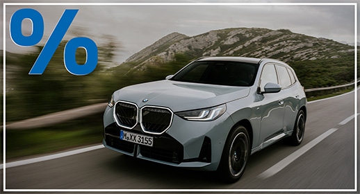 Hellgrauer BMW X3 SUV auf einer kurvigen Küstenstraße bei bewölktem Himmel; blaues Prozent-Symbol oben links.