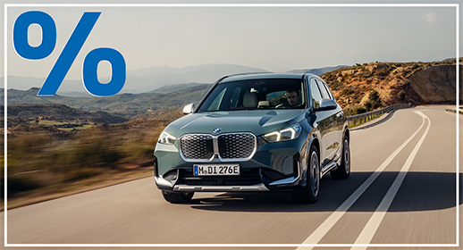 Graugrüner BMW iX1 Elektro-SUV auf einer hellen Landstraße unter klarem Himmel; blaues Prozent-Symbol oben links.