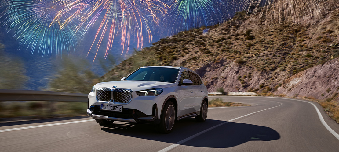 Das Bild zeigt einen weißen, vollelektrischen BMW iX1, der eine sonnige Passstraße in den Bergen befahren hat. Die Grafik nutzt die gleichen Kampagnenelemente: Ein großes blaues Prozentzeichen (%) oben links und funkelnde Feuerwerkseffekte am oberen Bildrand. Der weiße Rahmen unterstreicht den Charakter eines Sonderangebots. Die Szene kombiniert umweltbewusste Elektromobilität mit dem Versprechen attraktiver Ersparnisse zum Jahresbeginn.