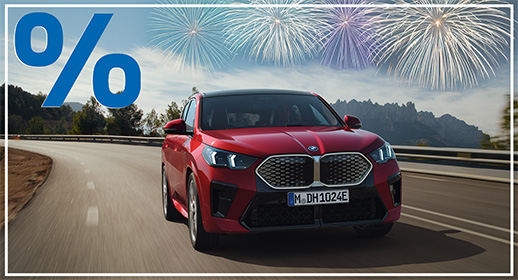 Diese Werbegrafik zeigt einen vollelektrischen BMW iX2 in einem leuchtenden Rot, der dynamisch eine kurvige Bergstraße hinauffährt. Das SUV-Coupé ist frontal aus einer tiefen Perspektive abgebildet, was seine sportliche Front mit dem markanten Grill und den modernen Scheinwerfern betont. Im Hintergrund sind bewaldete Hänge und schroffe Felsformationen unter einem leicht bewölkten Himmel zu sehen.  Über der Bergkulisse erstrahlen mehrere farbenfrohe Feuerwerke in Gold-, Blau- und Violetttönen. Das einheitliche Design der Serie wird auch hier durch das große blaue Prozentzeichen in der oberen linken Ecke und den schmalen weißen Rahmen vervollständigt.