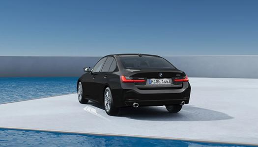 BMW 3er Limousine Aktionsangebot Ausstattung Schwarze BMW 318i Limousine von hinten schräg dargestellt, auf einer weißen Plattform neben einem Wasserbecken unter blauem Himmel. Das Bild zeigt das Heck des spezifischen Modells des Angebots.