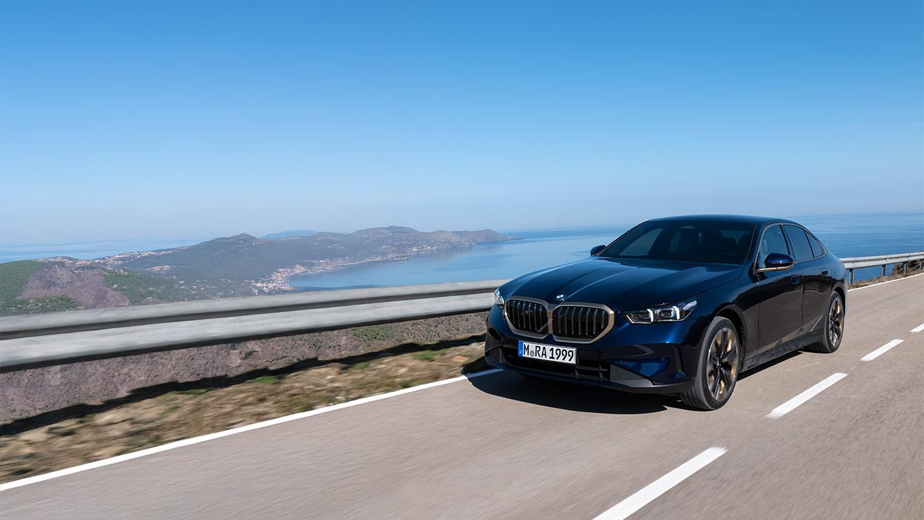 Eine dunkelblaue BMW 5er Limousine fährt auf einer Küstenstraße bei klarem, blauem Himmel mit weitem Ausblick auf das Meer und eine Hügellandschaft im Hintergrund.