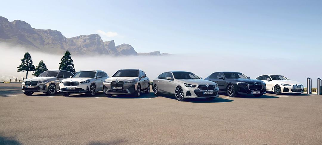 Eine Reihe von sechs verschiedenen weißen, silbernen und dunklen BMW Elektroautos steht nebeneinander auf einem Parkplatz im Freien, mit einer bergigen Landschaft im Hintergrund.