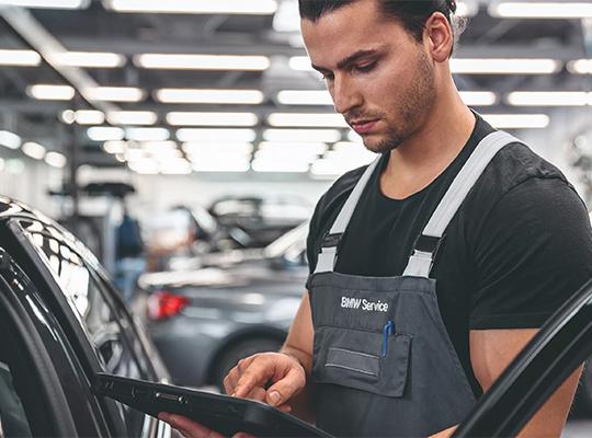 Ein Mechaniker in BMW Service-Arbeitskleidung beugt sich über ein Auto in einer Werkstatt und bedient ein Diagnosegerät oder Tablet.