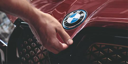 Eine Nahaufnahme zeigt die rote Motorhaube eines BMW Elektroautos. Eine Hand berührt die Kante der Haube direkt neben dem blau-weißen BMW-Logo, das von einem blauen Ring umgeben ist. Darunter ist ein Teil des charakteristischen Kühlergrills mit dreieckiger Musterung zu sehen.
