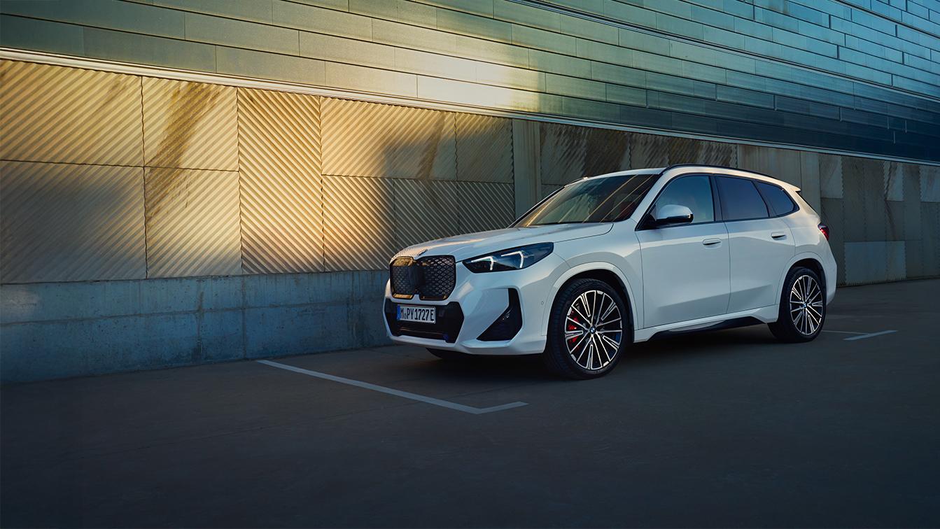 Weißer BMW iX1 in Front-Seitenansicht vor einer modernen Gebäudefassade bei warmem Sonnenlicht