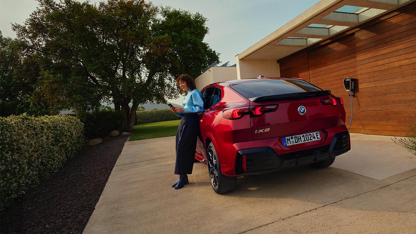 Roter BMW iX2 in Heckansicht vor einem modernen Wohnhaus mit Ladestation. Eine Frau steht neben dem Fahrzeug.