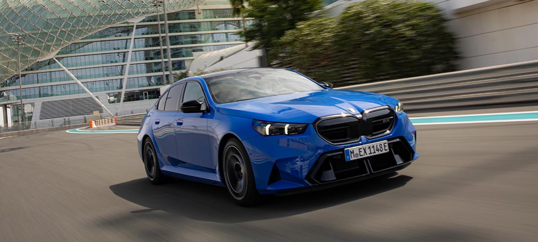 In der Bildmitte ist eine blaue BMW M5 Limousine in Bewegung zu sehen. Das Auto fährt auf einer Rennstrecke. Das Gebäude im Hintergrund hat eine besondere, futuristische Architektur. Die Beleuchtung ist hell, und das Bild wurde vermutlich tagsüber aufgenommen.
