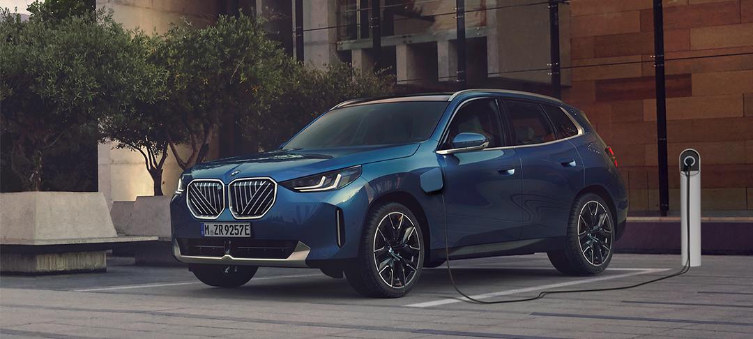 Das Bild zeigt ein Auto, einen BMW X3 Plug-in-Hybrid, das in einem städtischen Umfeld geparkt und an eine Ladesäule angeschlossen ist. Es ist ein blau-metallicfarbenes SUV, das von vorne links zu sehen ist. Die Front des Autos ist geprägt von den markentypischen doppelten Nieren, die hier in einem auffälligen, silbernen Gitterdesign gehalten sind. Links von der Fahrertür ist der Ladeanschluss sichtbar, von dem ein schwarzes Kabel zu einer weißen Ladesäule rechts vom Auto führt. Das Auto steht auf einem gepflasterten Platz neben einem kleinen Baum und einer modernen Sitzbank. Der Hintergrund besteht aus einem Gebäude mit einer strukturierten Fassade. Das Licht ist eher gedämpft, was dem Bild eine ruhige und elegante Atmosphäre verleiht.