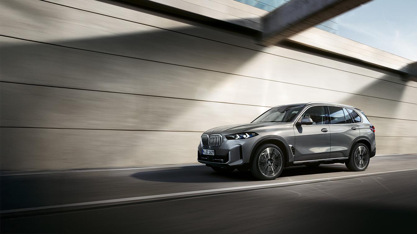 Das Bild präsentiert einen silber-grauen BMW X5 in einer dynamischen Fahraufnahme vor einer minimalistischen Betonkulisse. Durch die gezielte Bewegungsunschärfe der Räder und des Bodens wird ein Eindruck von Geschwindigkeit und Eleganz vermittelt, während die harten Schattenwürfe der Architektur das moderne Design des SUVs unterstreichen. Die kühle Lichtstimmung und die hochwertige Optik des Fahrzeugs verleihen der Szene einen exklusiven, technologisch orientierten Charakter, der typisch für ein professionelles Automobil-Leasingangebot ist.