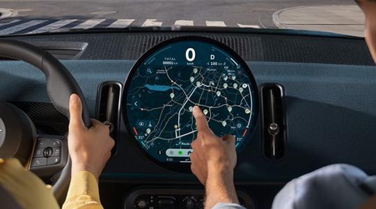 Das achte Bild zeigt den MINI Routenplaner auf dem kreisförmigen OLED-Display, der speziell für die Elektromobilität entwickelt wurde. Das System berechnet automatisch ladeoptimierte Routen, indem es notwendige Ladestopps basierend auf der aktuellen Reichweite und Echtzeit-Verkehrsdaten in die Zielführung integriert. Die übersichtliche Darstellung auf der Karte zeigt dem Fahrer nicht nur den Weg, sondern auch die voraussichtliche Ankunftszeit inklusive der benötigten Ladezeit an. Diese intelligente Vernetzung sorgt dafür, dass auch längere Strecken mit den elektrischen MINI-Modellen komfortabel und ohne Reichweitenangst bewältigt werden können.