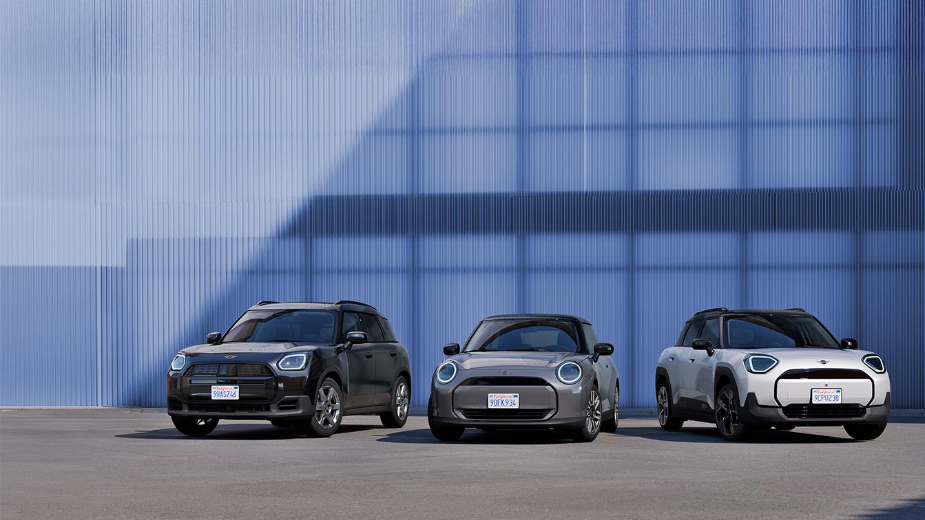 Drei Modelle der neuen MINI-Familie präsentieren sich vor einer blau schimmernden, industriellen Kulisse: Der schwarze Countryman E, der klassisch graue Cooper E und der weiße Aceman. Das minimalistische Design der Fahrzeuge und die kühle Architektur im Hintergrund unterstreichen den modernen, vollelektrischen Charakter der aktuellen Modellpalette.