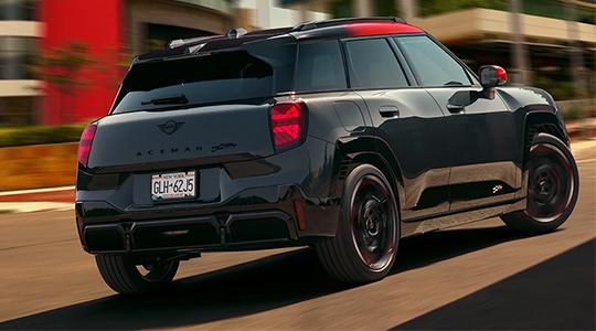 Das fünfte Bild zeigt den MINI John Cooper Works (JCW) Aceman Electric, die sportliche Speerspitze der Modellreihe. In einer dynamischen Heckansicht bei voller Fahrt wird der Fokus auf das exklusive JCW-Design gelenkt, das sich durch ein kontrastreiches rotes Dach und rote Außenspiegelkappen von den Standardmodellen abhebt. Schwarze Aerodynamik-Elemente wie der Heckdiffusor und die markanten Winglets unterstreichen den Rennsport-Charakter und optimieren den Luftwiderstand. Mit einer gesteigerten Leistung von 258 PS (190 kW) sprintet dieser elektrische Crossover in nur 6,4 Sekunden von 0 auf 100 km/h. Das Bild fängt perfekt das für MINI typische „Gokart-Feeling“ ein, das hier durch sofort verfügbares Drehmoment und ein speziell abgestimmtes Fahrwerk neu definiert wird.