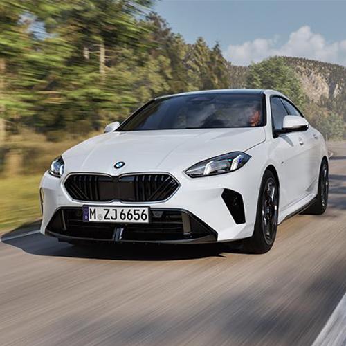 Es handelt sich um ein weißes BMW 2er Gran Coupé, das dynamisch auf einer kurvigen Landstraße unterwegs ist. Das Auto ist in der Mitte des Bildes von vorne zu sehen. Der Kühlergrill ist schwarz, und die LED-Scheinwerfer sind scharf gezeichnet. Im Hintergrund sind grüne Bäume und Berge unter einem hellen Himmel. Das Bild ist so aufgenommen, dass es Geschwindigkeit vermittelt, was durch die leichte Unschärfe des Hintergrundes noch verstärkt wird. Das Kennzeichen lautet "M ZJ 6656".