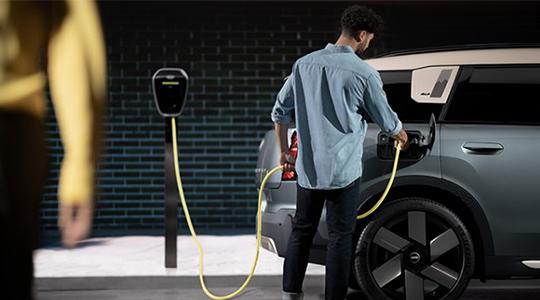 In dieser Szene steht der Ladevorgang eines modernen Elektroautos im Mittelpunkt. Ein Mann, bekleidet mit einem hellblauen Jeanshemd und einer dunklen Hose, ist gerade dabei, ein auffälliges gelbes Ladekabel in den rückwärtigen Anschluss eines Fahrzeugs zu stecken. Das Auto, ein SUV in einem eleganten Grau-Blau mit kontrastierendem cremeweißem Dach, ist durch das Kabel direkt mit einer schlanken, schwarzen Ladestation verbunden, die auf einer Stele montiert ist.  Die Umgebung wirkt wie eine moderne Garage oder ein Parkdeck bei Nacht, was durch die dunkle Backsteinwand im Hintergrund und die gezielte Beleuchtung unterstrichen wird. Besonders markant sind die großen, aerodynamisch gestalteten Felgen des Wagens, die dem Fahrzeug ein futuristisches Aussehen verleihen. Eine unscharfe Silhouette am linken Bildrand erzeugt eine Tiefenwirkung, die den Blick des Betrachters unmittelbar auf die Interaktion zwischen Mensch, Technik und Infrastruktur lenkt.