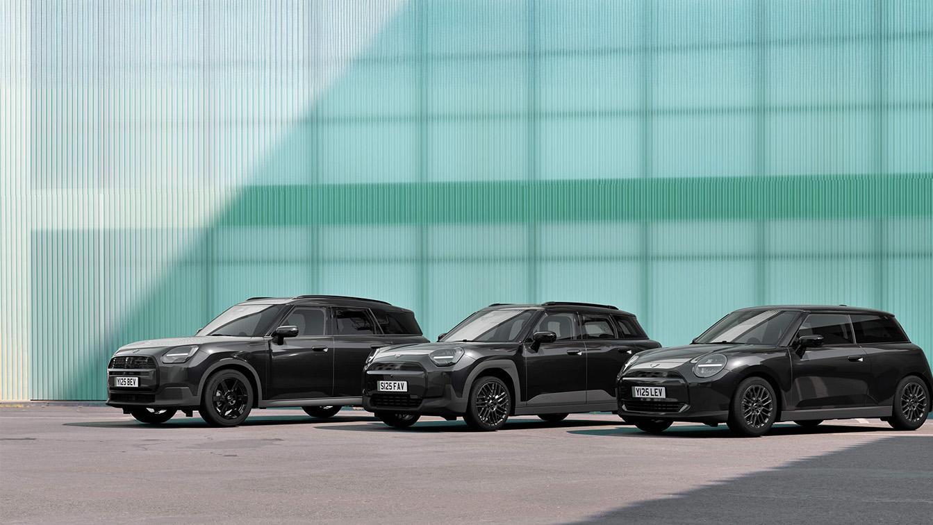 Das Bild zeigt drei verschiedene Modelle der neuen MINI-Generation in einer eleganten, komplett schwarzen Sonderedition. Die Fahrzeuge – ein großer Countryman, ein kompakter Aceman und ein klassischer Dreitürer – stehen nebeneinander auf einer grauen Asphaltfläche vor einer modernen Fassade aus helltürkisem Profilglas. Durch den „All-Black-Look“ wirken die Autos besonders hochwertig und urban, während die vertikalen Linien der hellen Glaswand im Hintergrund einen frischen, kühlen Kontrast bilden. Die Szenerie ist in helles Tageslicht getaucht, was die glänzenden Oberflächen der schwarzen Karosserien und die markanten Felgen betont.  