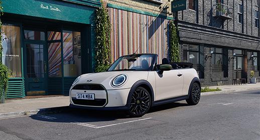 Ein cremeweißes MINI Cabrio der limitierten Paul Smith Edition mit geöffnetem Verdeck, geparkt vor einem Paul Smith Store mit einer bunt gestreiften Fassade.