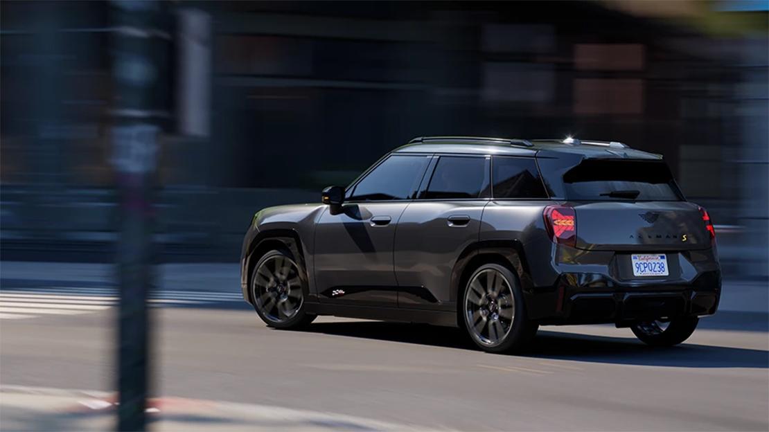 Bild zeigt den MINI Aceman Electric in einer dynamischen Fahraufnahme bei Dämmerung oder in einer schattigen städtischen Umgebung. Das Fahrzeug ist in einem edlen, dunklen Graumetallic gehalten, wobei das Heckdesign mit den charakteristischen, vertikalen LED-Rückleuchten im Union-Jack-Stil besonders betont wird. Die Bewegungsunschärfe des Hintergrunds vermittelt ein Gefühl von Geschwindigkeit und unterstreicht das für MINI typische agile Fahrverhalten. Als kompaktes Crossover-Modell bietet er trotz seiner urbanen Maße Platz für fünf Personen und kombiniert moderne Elektromobilität mit einem sportlichen Auftritt.