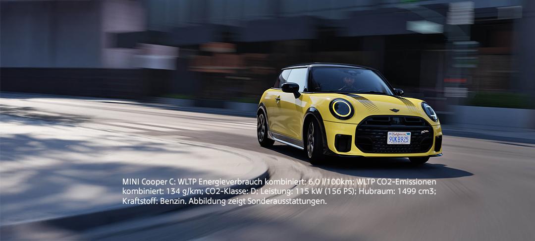 Ein gelber Mini Cooper C mit schwarzen Akzenten und schwarzen Felgen fährt dynamisch auf einer Straße mit leichtem Bewegungsunschärfe-Effekt im Hintergrund, der die Geschwindigkeit andeutet. Das Auto hat zwei schwarze Streifen auf der Motorhaube und runde Scheinwerfer. Im unteren Bereich des Bildes sind technische Daten des MINI Cooper C auf Deutsch aufgelistet: WLTP Energieverbrauch kombiniert: 6,0 l/100km; WLTP CO2-Emissionen kombiniert: 134 g/km; CO2-Klasse: D; Leistung: 115 kW (156 PS); Hubraum: 1499 cm3; Kraftstoff: Benzin. Es wird auch erwähnt, dass die Abbildung Sonderausstattungen zeigt.