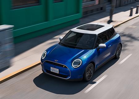 Lagerwagenaktion MINI BMW Rhein Ein blauer Mini Cooper auf einer Straße in einer städtischen Umgebung. Das Auto hat ein weißes Dach und die typischen runden Mini-Scheinwerfer mit einer modernen Lichtsignatur. Es fährt auf einer Straße, die von einem Gehweg und Gebäuden gesäumt ist, darunter ein Gebäude mit einer grünen Fassade auf der linken Seite. Die Perspektive ist leicht von oben und schräg von vorne.