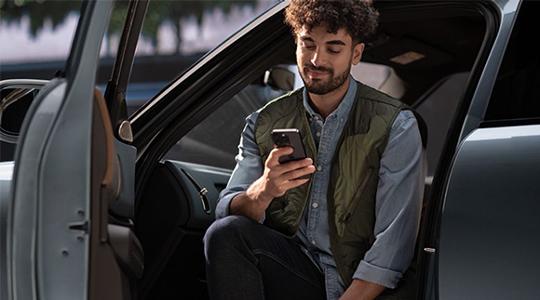  In diesem Bild sieht man einen jungen Mann mit lockigem Haar und einem Bart, der entspannt auf dem Fahrersitz eines Autos sitzt, während die Fahrertür weit offen steht. Er trägt ein hellblaues Hemd unter einer olivgrünen Steppweste und blickt mit einem leichten Lächeln auf sein Smartphone, das er mit beiden Händen hält.  Die Perspektive ist leicht von außen durch die offene Türöffnung gewählt, was den Blick auf das moderne und schlichte Interieur des Wagens freigibt. Das Licht fällt weich von der Seite ein und erzeugt eine ruhige, alltägliche Atmosphäre. Im Hintergrund sind vage die Umrisse von Bäumen oder Sträuchern zu erkennen, was darauf hindeutet, dass das Fahrzeug im Freien geparkt ist.
