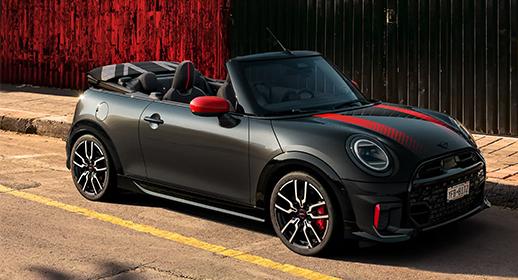 Ein glänzendes, dunkelgraues MINI John Cooper Works Cabriolet mit geöffnetem schwarzen Stoffverdeck, roten Spiegelkappen und roten Rennstreifen auf der Motorhaube. Das Fahrzeug steht auf einem Bürgersteig vor einer roten Wand und hat sportliche schwarze Felgen mit roten Bremssätteln.