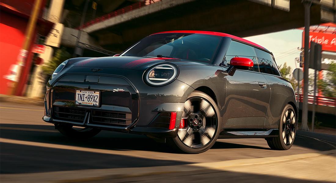 Ein glänzender, dunkelgrauer MINI John Cooper Works Electric mit rotem Dach, roten Spiegelkappen und roten Akzenten an der Front. Das Auto ist in Bewegung auf einer Straße bei sonnigem Wetter, mit verschwommenem Hintergrund, der urbane Elemente andeutet.