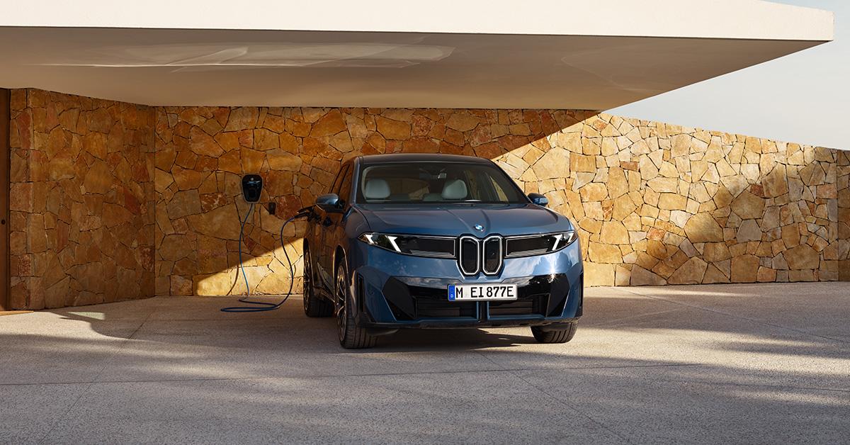 Dieses Bild zeigt die Front eines dunkelblauen Elektrofahrzeugs in einer luxuriösen, steinernen Umgebung. Das Auto ist unter einem hellen Vordach geparkt. Das Ladekabel ist mit einer Wallbox an der Wand verbunden. Die Wand ist das auffälligste Element und besteht aus unregelmäßig geschichteten, goldbraunen Natursteinen. Das kräftige Licht der tief stehenden Sonne wirft lange Schatten und betont die Textur der Steinwand, was der gesamten Szene einen sehr exklusiven und mediterranen Charakter verleiht.