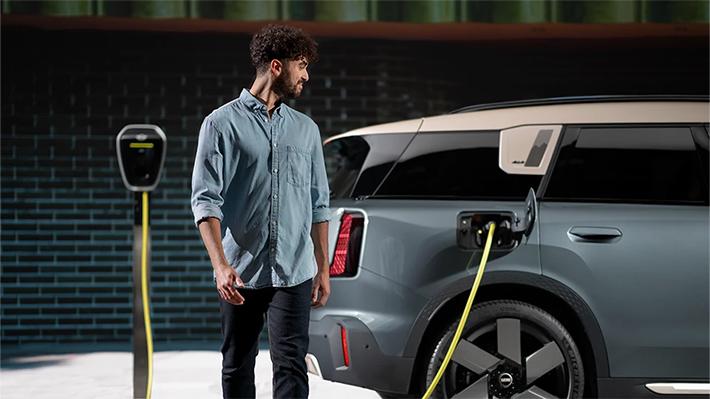 In dieser modernen, urbanen Aufnahme steht die Elektromobilität im Mittelpunkt. Auf der rechten Seite ist das Heck eines markanten SUVs zu sehen, der durch seine zweifarbige Gestaltung in mattem Blaugrau und einem beigefarbenen Dach auffällt. Das Fahrzeug ist über ein leuchtend gelbes Kabel mit einer schlanken, schwarzen Ladestation verbunden, die im linken Hintergrund platziert ist.  Ein junger Mann mit lockigem Haar, bekleidet mit einem hellblauen Jeanshemd und dunkler Hose, geht gerade an dem Wagen vorbei und blickt dabei aufmerksam über seine Schulter zurück zum Ladevorgang. Die Szene spielt sich vor einer dunklen, strukturierten Ziegelwand ab, die dem Bild einen industriellen und hochwertigen Charakter verleiht. Das Zusammenspiel aus dem punktuellen Licht und dem kräftigen Gelb des Kabels setzt einen modernen Akzent und unterstreicht den technologischen Fortschritt im Alltag.