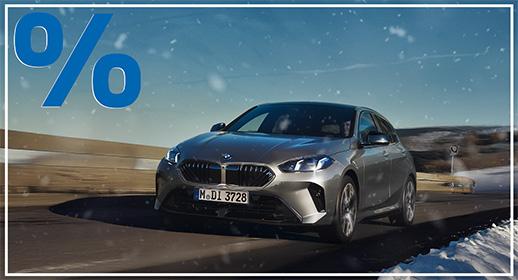 Das Bild zeigt einen silber-grauen BMW 1er in einer winterlichen Szene. Das Auto fährt auf einer verschneiten oder zumindest winterlich wirkenden Straße oder Brücke. Im Hintergrund sind Berge oder eine hügelige Landschaft zu sehen. Es schneit leicht und die Gesamtstimmung ist kühl und klar. In der oberen linken Ecke des Bildes ist prominent ein großes, blaues Prozentzeichen (%) platziert, was auf ein Sonderangebot oder eine Werbeaktion hindeutet.