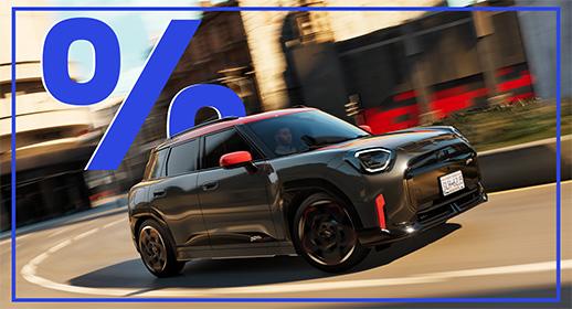 Das Bild zeigt einen grauen MINI Aceman im John Cooper Works (JCW) Design. Das Auto hat ein rotes Dach, rote Außenspiegel und rote Akzente an der Front. Es fährt auf einer kurvigen Straße in einer städtischen Umgebung. Im oberen linken Bereich des Bildes ist ein großes, blaues Prozentzeichen (%) zu sehen. Das Kennzeichen lautet "D81-43B".