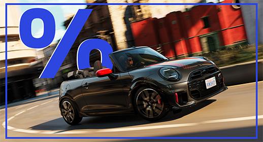 Das Bild zeigt ein graues MINI Cabriolet im John Cooper Works (JCW) Design. Das Auto hat ein offenes Verdeck und rote Außenspiegel. Es fährt auf einer kurvigen Straße in einer städtischen Umgebung. Im oberen linken Teil des Bildes schwebt ein großes, blaues Prozentzeichen (%). Das Kennzeichen lautet "D 9L 389".