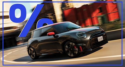 Das Bild zeigt einen grauen MINI Cooper Elektro im John Cooper Works (JCW) Design. Das Auto hat rote Außenspiegel und ein rotes Dach, sowie rote Akzente an der Front. Das Auto fährt auf einer Straße, und im oberen linken Teil des Bildes schwebt ein großes, blaues Prozentzeichen (%). Das Kennzeichen lautet "0K2-882".