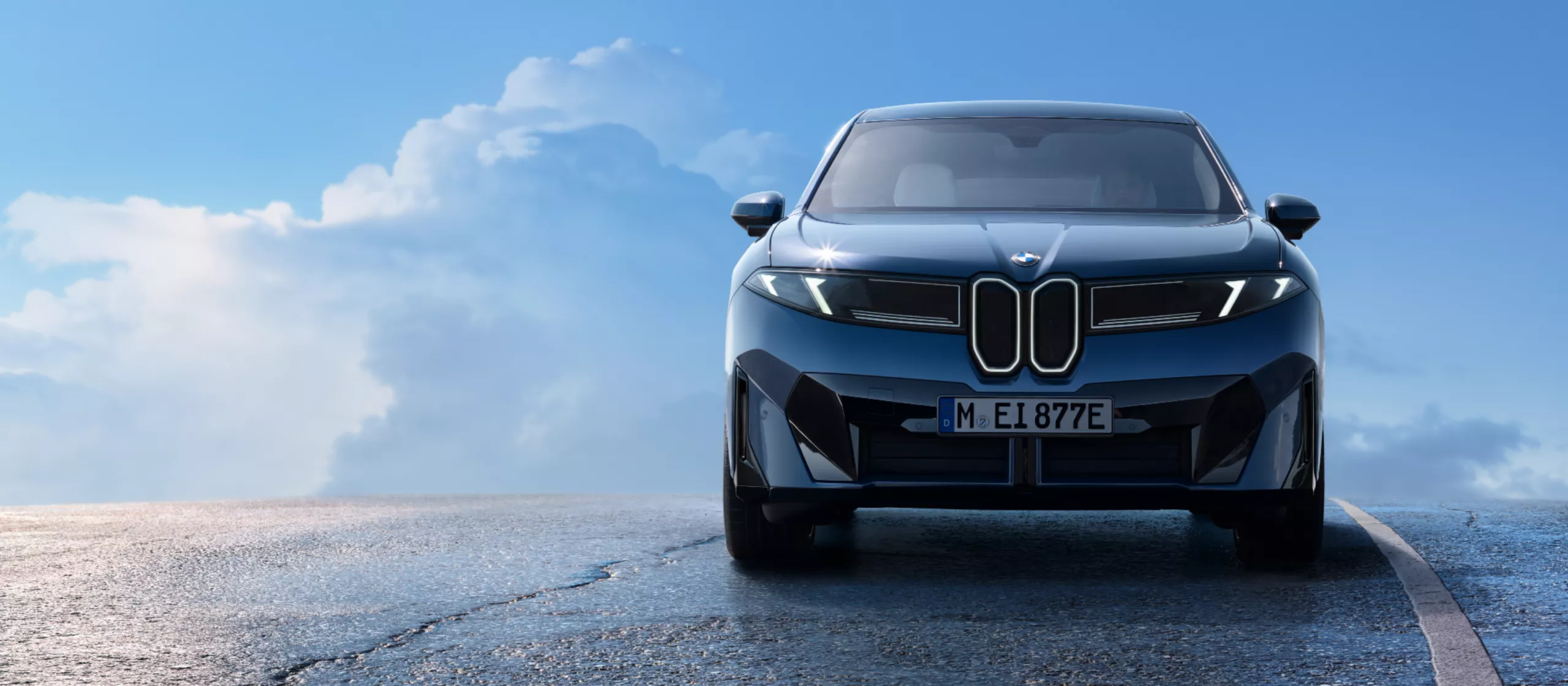 Das Bild zeigt einen blauen BMW iX von vorne auf einer nassen, asphaltierten Straße. Der Himmel im Hintergrund ist blau mit weißen Wolken. Das Fahrzeug ist der Hauptfokus des Bildes und die Frontansicht ist gut sichtbar.