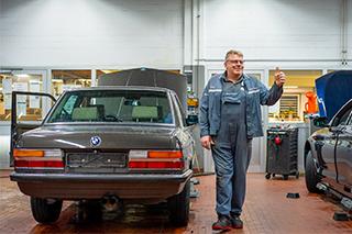 Ein lächelnder Mechaniker in blauer Arbeitskleidung steht in einer Werkstatt neben einem älteren BMW-Modell mit geöffneter Motorhaube und zeigt einen Daumen hoch.